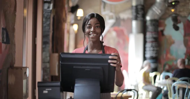Positive young woman in uniform smiling while standing at counter desk in cafe