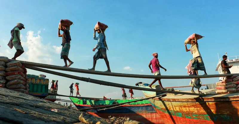 Laborers balancing bags on planks by colorful boats under clear blue skies.