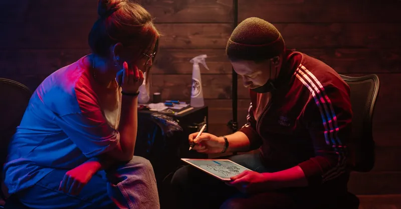 Tattoo artist sketching designs on tablet in dimly lit studio, creating unique artwork.
