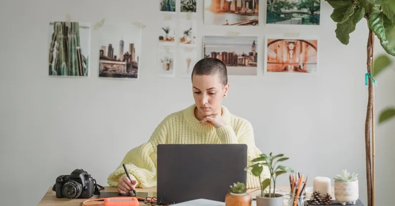 Female graphic designer working on a laptop with a stylus in a cozy home office filled with plants and art.