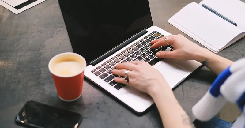 A freelancer typing on a laptop with a cup of coffee, smartphone, and notepad on the table.