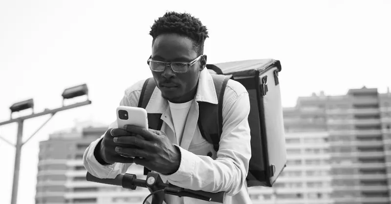 Black and white image of a delivery rider leaning on scooter checking smartphone.