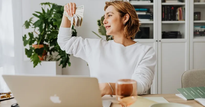 Smiling woman holding cash, working at home office with a laptop, symbolizing financial success.