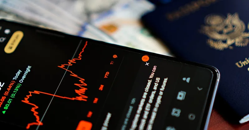 Close-up of a smartphone displaying a stock market app alongside a US passport and currency.