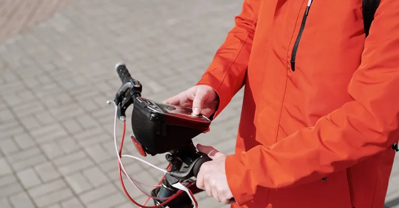 A food delivery cyclist in an orange jacket uses a smartphone while riding in the city.