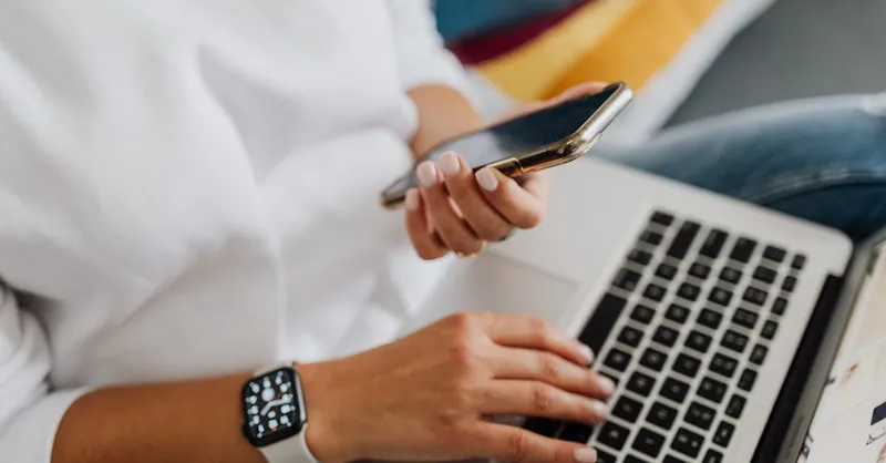 Close-up of a person using a smartphone and laptop for work, showcasing multitasking in a modern home office setting.