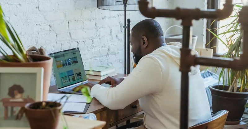 Side view of concentrated African American male freelancer sitting at wooden table and working online on netbook