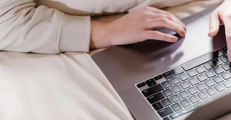 A woman relaxes and works on her laptop in a cozy bedroom environment, showing remote work lifestyle.