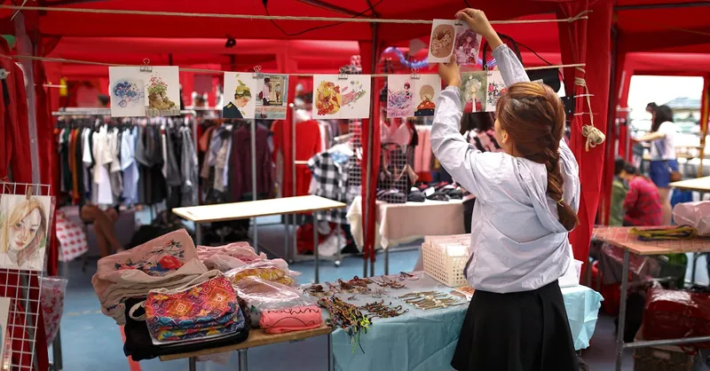 A woman organizes art prints in a vibrant outdoor market stall with colorful clothing and accessories.