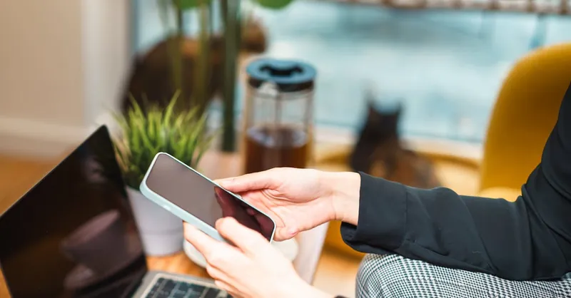 Woman working remotely from home, using a smartphone and laptop at a cozy desk with a plant and cat in background.