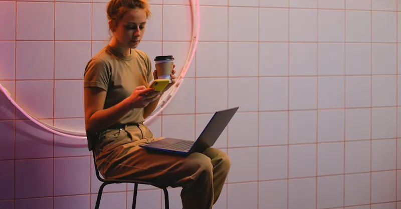Young woman multitasks with phone and laptop in a trendy cafe with neon lighting.