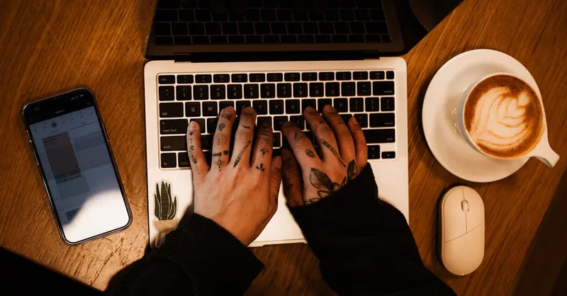 Tattooed hands on a laptop with coffee and phone, perfect workspace inspiration.