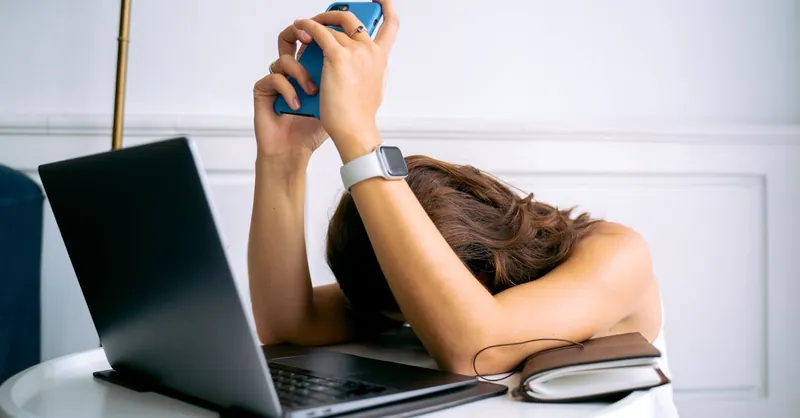 A woman overwhelmed by work, resting head on table with laptop, phone, and smartwatch.