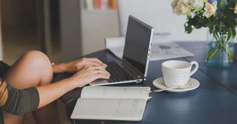 Female freelancer using laptop with coffee at home office desk, surrounded by roses and a planner.